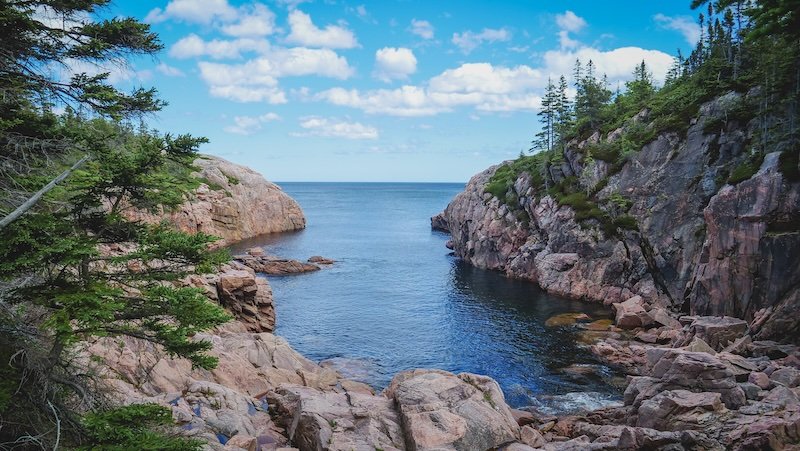 Dramatic cliffs of pink granite frame a narrow cove opening into the Atlantic Ocean, surrounded by evergreen forest and blue skies along the rugged coastline of the Cabot Trail in Cape Breton Highlands National Park, Nova Scotia