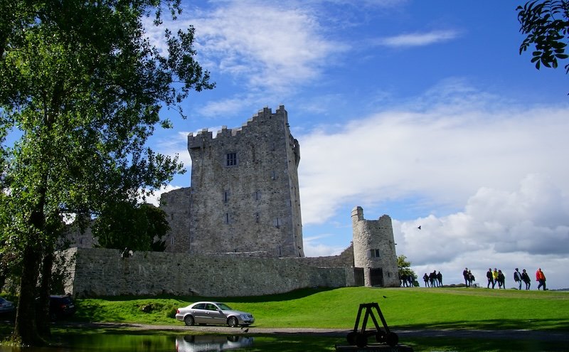 Stone fortress of Ross Castle standing on the lush green shores of Killarney National Park in County Kerry, Ireland, with visitors walking along the grassy grounds under a bright blue sky dotted with clouds during a scenic road trip through Ireland