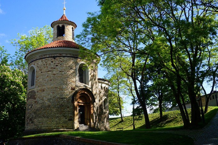 Historic Rotunda of St. Martin with its circular stone walls and red-tiled roof surrounded by lush green trees in Vyšehrad fortress park in Prague, Czech Republic, on a sunny afternoon