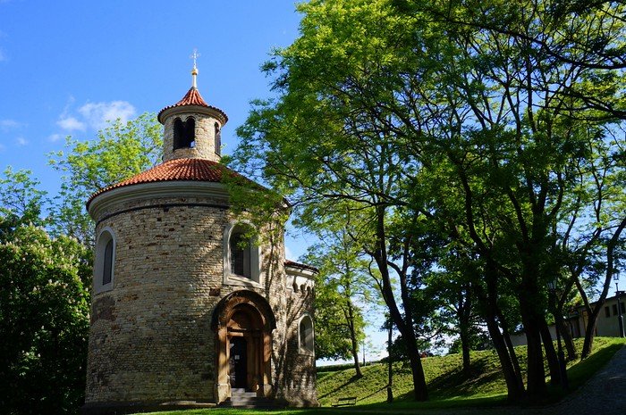 Rotunda of Saint Martin in Prague, Czech Republic, a historic 11th-century stone church with a red-tiled roof and small tower, surrounded by leafy green trees under a bright blue sky, located on the Vyšehrad hill