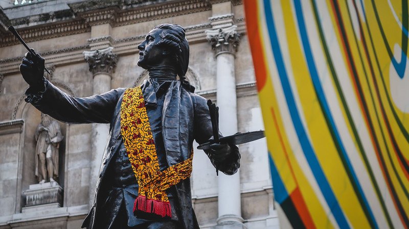 Statue outside the Royal Academy of Art in London adorned with a colorful patterned scarf, set against the building’s grand classical architecture and vibrant contemporary art installation, blending history with modern creativity in the city
