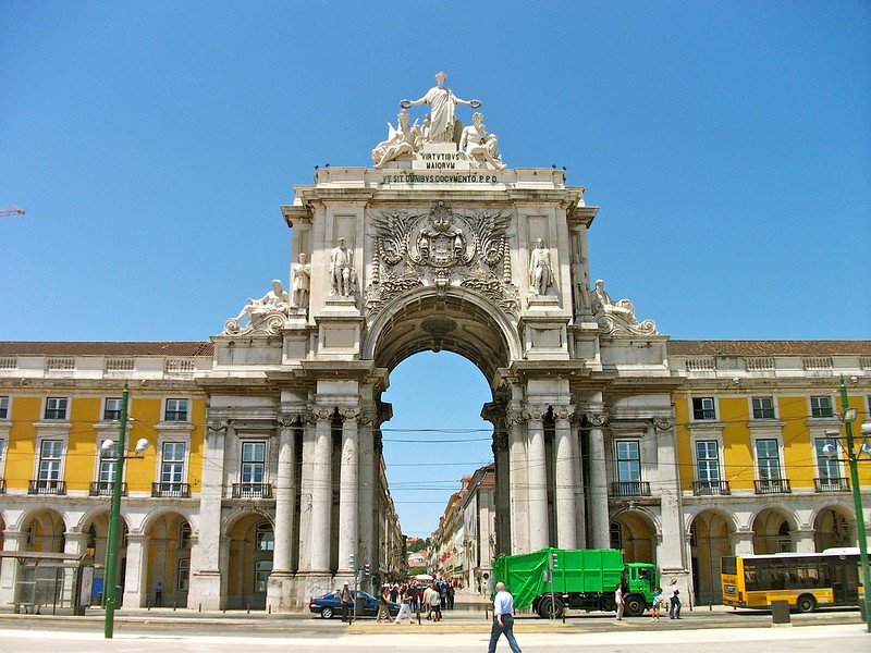 The Rua Augusta Arch in Lisbon, Portugal, featuring ornate sculptures, detailed stone carvings, and a grand central archway with yellow buildings on each side under a bright blue sky, marking the entrance to the city’s downtown area