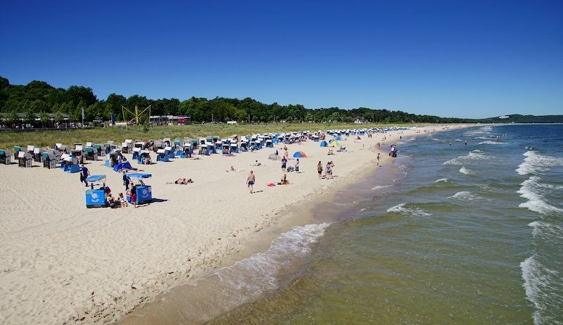 Sunny summer day at a sandy beach on Rügen Island, Germany, with rows of colorful beach chairs (strandkorb), sunbathers, and gentle waves from the Baltic Sea rolling onto the shore