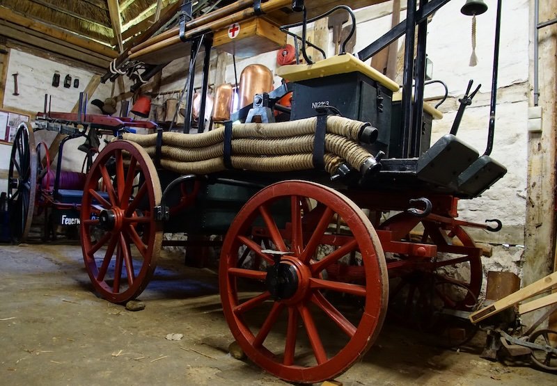Historic horse-drawn fire engine on display at a museum on Rügen Island, Germany, featuring red wooden wheels, coiled hoses, and copper water tanks inside a rustic thatched-roof barn