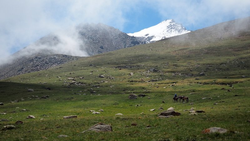 A lone horse rider leading pack horses across a remote grassy alpine slope dotted with rocks, with mist swirling around rugged peaks and a snow-capped summit in the background during the 3-Day Panorama Trek in Kyrgyzstan
