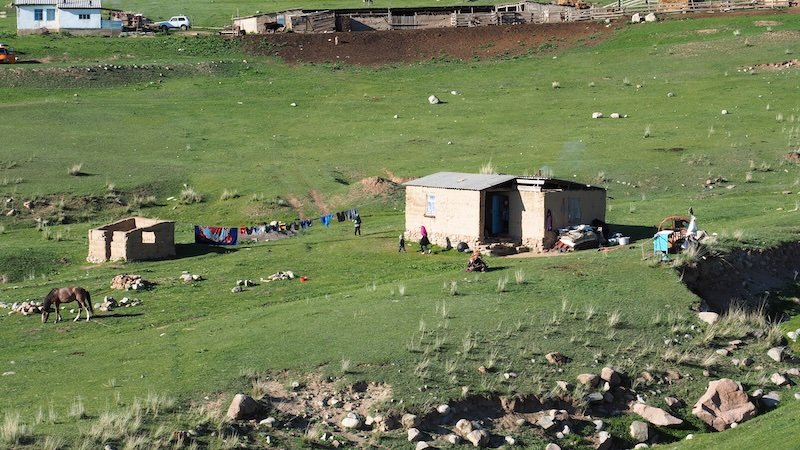 A small rural home in the green hills of Kyrgyzstan with laundry hanging on a clothesline, children playing outside, and a horse grazing nearby during the 3-Day Panorama Trek