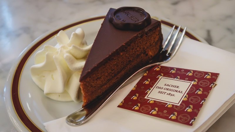 Close-up of a decadent slice of Sachertorte served with whipped cream on a china plate at Vienna’s historic Café Sacher, showcasing its glossy chocolate glaze and signature chocolate seal Close-up of a decadent slice of Sachertorte served with whipped cream on a china plate at Vienna’s historic Café Sacher, showcasing its glossy chocolate glaze and signature chocolate seal