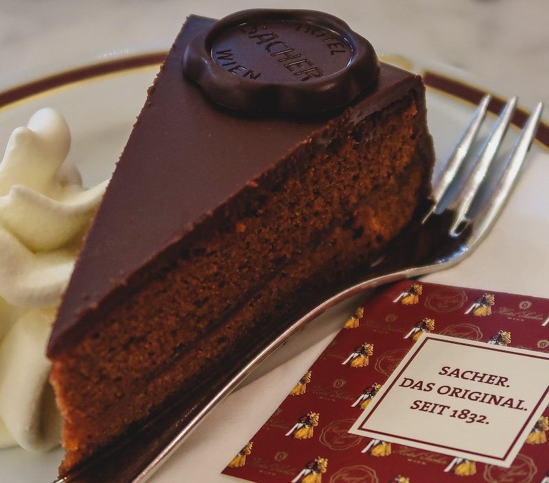 Close-up of a decadent slice of Sachertorte served with whipped cream at the historic Hotel Sacher in Vienna, showcasing its glossy chocolate glaze and signature chocolate seal Close-up of a decadent slice of Sachertorte served with whipped cream at the historic Hotel Sacher in Vienna, showcasing its glossy chocolate glaze and signature chocolate seal