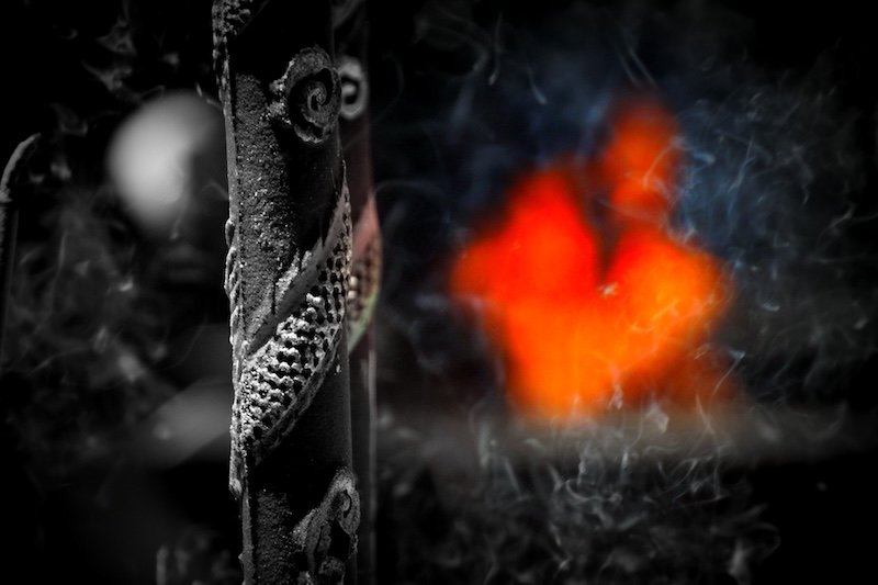 Close-up of a carved incense stick holder with intricate dragon patterns releasing delicate swirls of smoke inside a dimly lit temple in Saigon (Ho Chi Minh City), Vietnam, with the warm orange glow of a ceremonial flame softly illuminating the background