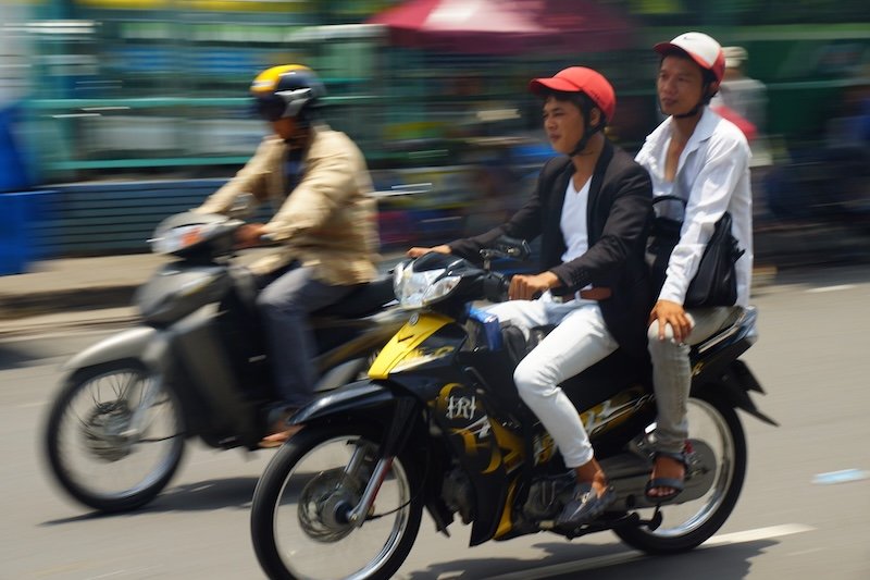 Motion-blurred street scene in Saigon (Ho Chi Minh City), Vietnam, showing several local riders zipping through city traffic on scooters and motorcycles, including two young men on a yellow motorbike wearing casual clothes and helmets as they weave through the bustling urban streets