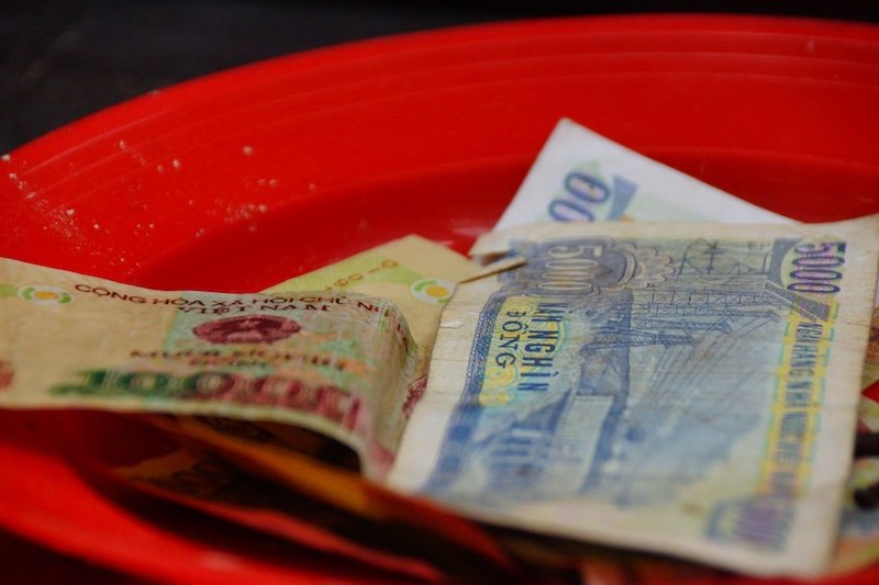 Close-up of Vietnamese dong banknotes placed in a red plastic dish in Saigon (Ho Chi Minh City), Vietnam, showing various denominations including worn and colorful bills with intricate designs and text, commonly used as local currency during everyday market transactions