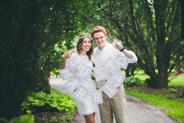 Samuel and Audrey Jeffery celebrate their wedding day by holding a “Just Married” banner while standing on a tree-lined path. Audrey beams in a short lace dress with a floral crown, while Samuel smiles proudly in a cream vest and shirt