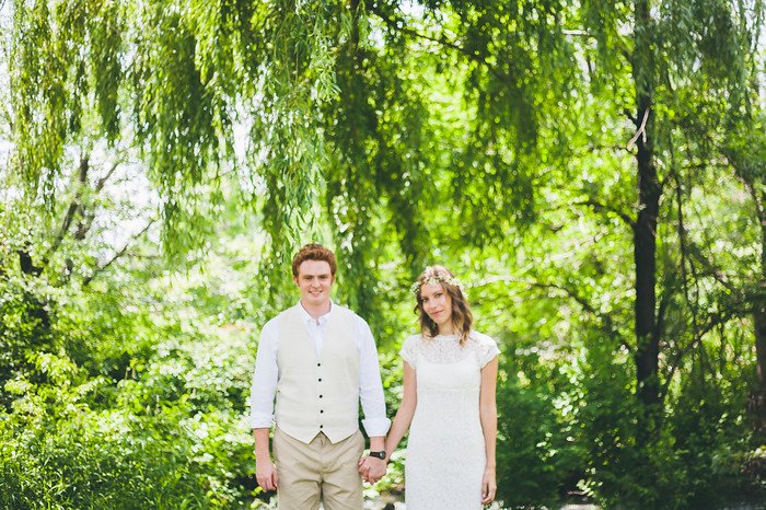 Samuel and Audrey Jeffery pose hand in hand as newlyweds beneath the lush green canopy of willow trees in Brampton, Ontario. Audrey wears a lace wedding dress with a floral crown while Samuel stands beside her in a cream vest and shirt, smiling proudly