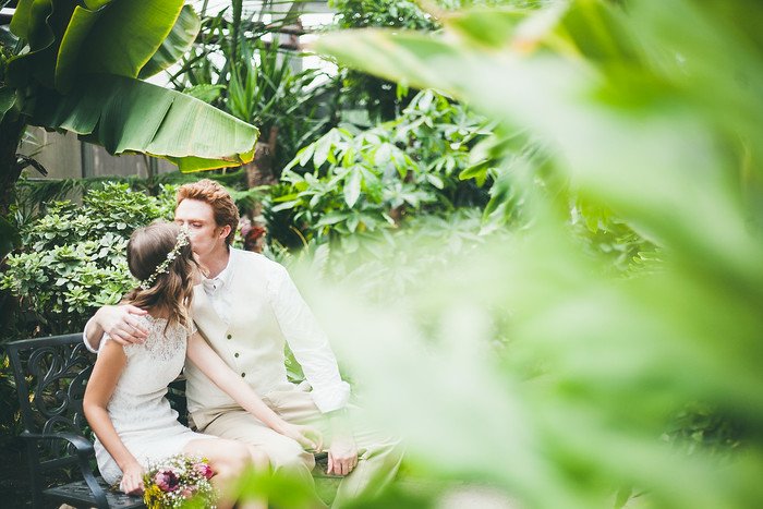 Newlyweds Samuel and Audrey share a romantic kiss on a garden bench surrounded by lush tropical greenery in Brampton, Ontario. Audrey wears a lace wedding dress with a floral crown while holding her bouquet, as Samuel embraces her in a cream vest