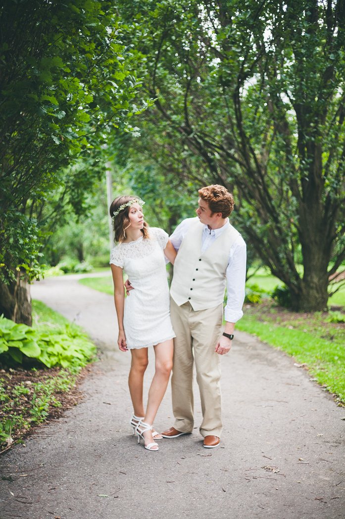 Samuel and Audrey share a playful moment on a tree-lined path during their summer wedding in Brampton, Ontario. Audrey wears a lace dress with a floral crown, while Samuel looks dapper in a vest and dress pants, capturing their fun-loving personalities