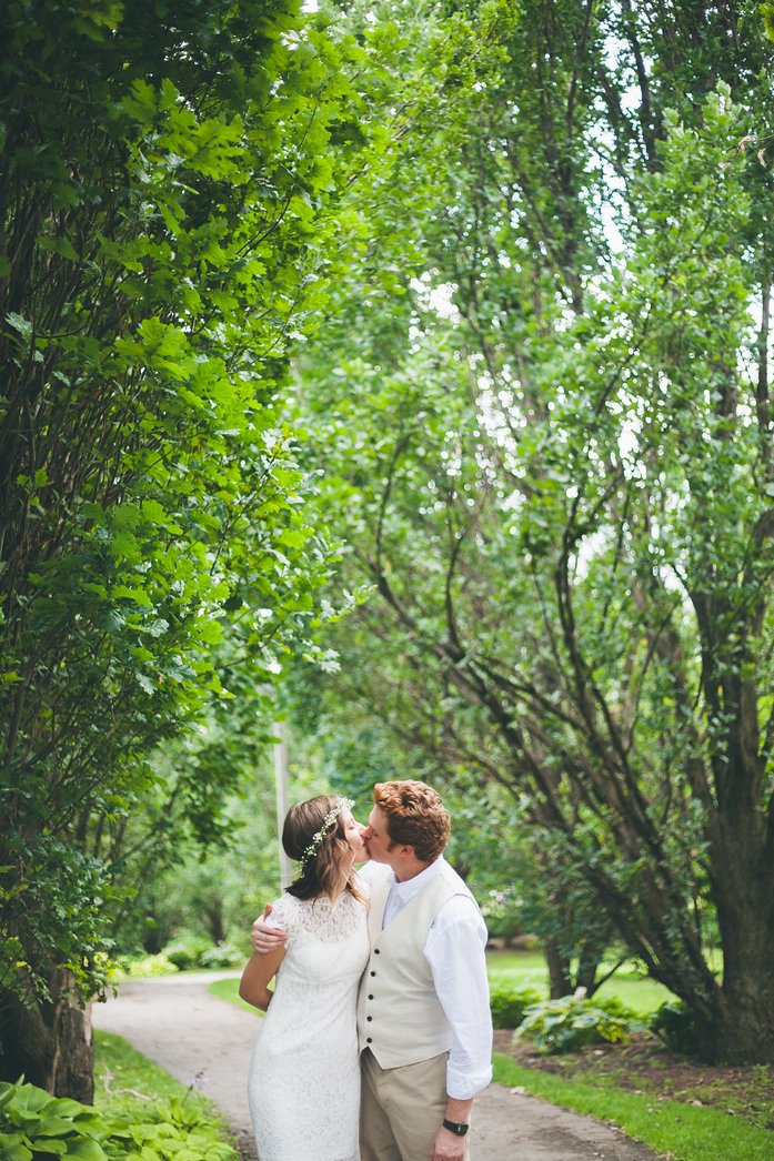 Samuel and Audrey share a tender kiss on a tree-lined path during their summer wedding in Brampton, Ontario. Audrey wears a lace dress with a delicate floral crown, while Samuel embraces her in a cream vest and shirt, capturing their love in nature
