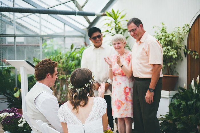 Samuel and Audrey seated together during their summer wedding ceremony in Brampton, Ontario, as Samuel’s parents stand alongside loved ones offering a heartfelt blessing. Surrounded by greenery in the tropical greenhouse, the moment feels intimate and meaningful