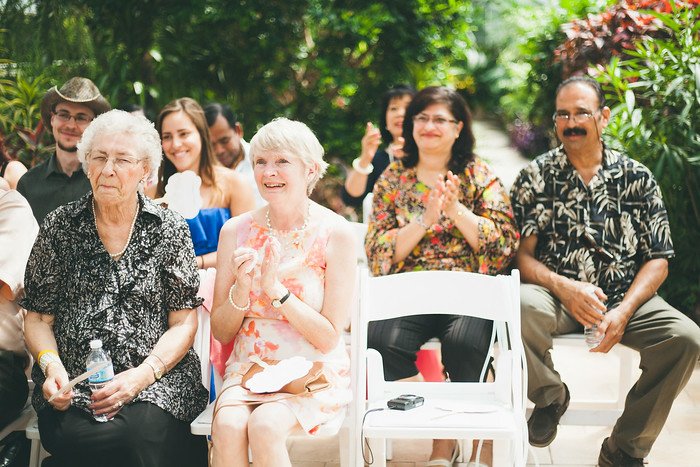Family and friends smile and applaud while attending Samuel and Audrey’s summer wedding ceremony in Brampton, Ontario. Guests are seated in white chairs inside a lush tropical greenhouse, enjoying the joyful atmosphere of the special celebration