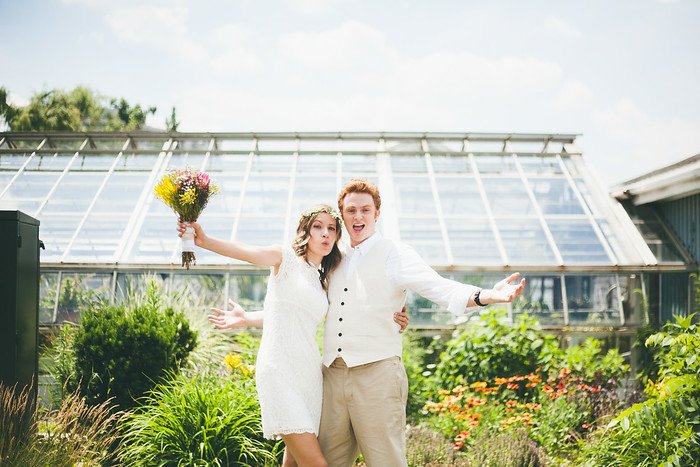Samuel and Audrey celebrating their summer wedding in a vibrant garden setting, posing joyfully in front of a greenhouse. Audrey wears a white lace dress with a flower crown and bouquet, while Samuel beams in a vest and slacks, both radiating happiness