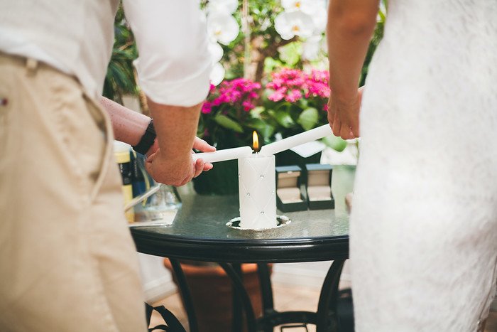Samuel and Audrey lighting a unity candle together during their summer wedding ceremony, symbolizing love and partnership. The intimate moment is set against a backdrop of vibrant flowers, adding warmth and romance to the celebration