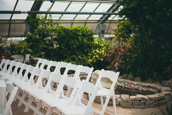 Rows of white chairs neatly arranged inside a lush tropical greenhouse, prepared for the summer wedding of Samuel and Audrey. The natural greenery, stone pond, and soft light create a serene, intimate, and romantic setting for the ceremony