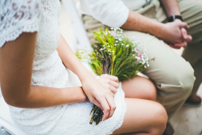 Close-up of Audrey in a delicate white lace wedding dress holding a rustic bouquet of wildflowers during her summer wedding with Samuel. Captured detail of hands and attire highlights the romantic simplicity and natural elegance of the celebration
