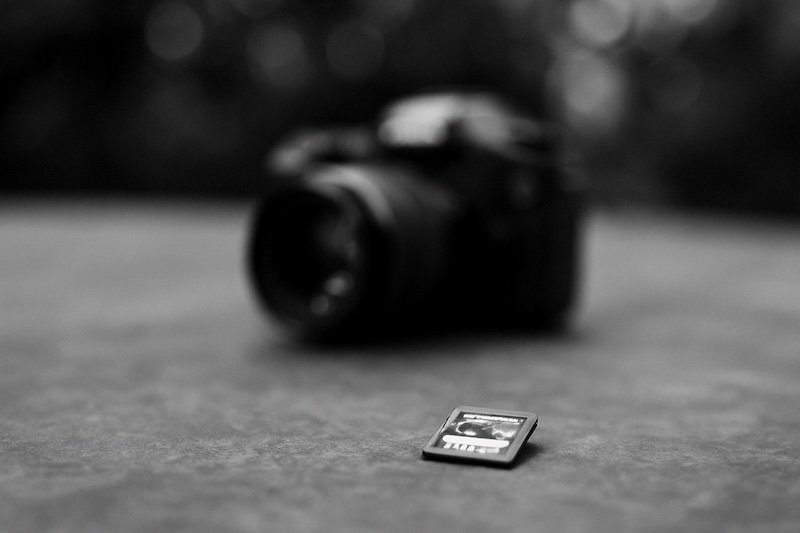 A black and white photograph of a SanDisk Extreme PRO 16GB memory card lying flat on a textured surface. In the distant background, a DSLR camera is softly blurred with strong bokeh, creating a cinematic, candid travel-gear aesthetic