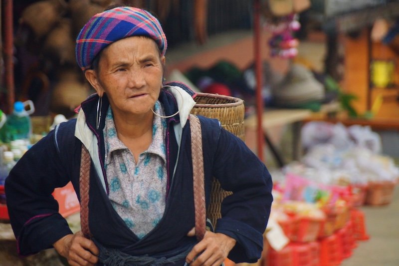 Elderly woman at a local market in Sapa, Vietnam wearing traditional hill-tribe clothing and carrying a woven basket backpack, surrounded by colorful market stalls and local goods