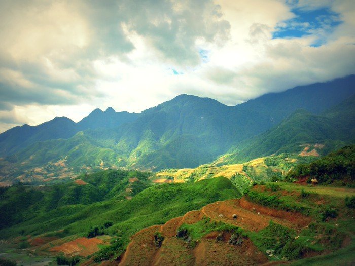 Panoramic view of lush green terraced hills and towering mountains under dramatic skies in Sapa, Vietnam, showcasing the region’s iconic rice paddies and breathtaking alpine scenery