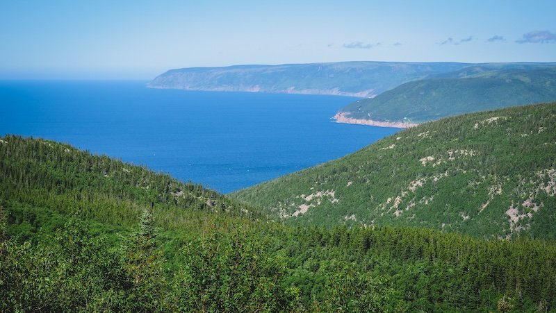 A sweeping view of forested green hills descending toward the deep blue waters of the Gulf of St. Lawrence, showcasing the dramatic coastal cliffs and natural beauty along the Cabot Trail in Cape Breton Highlands National Park, Nova Scotia
