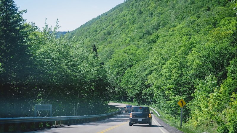 Cars wind along a curving two-lane highway surrounded by dense green forest and steep hillsides, capturing the lush scenery of a summer road trip along the Cabot Trail in Cape Breton Highlands National Park, Nova Scotia