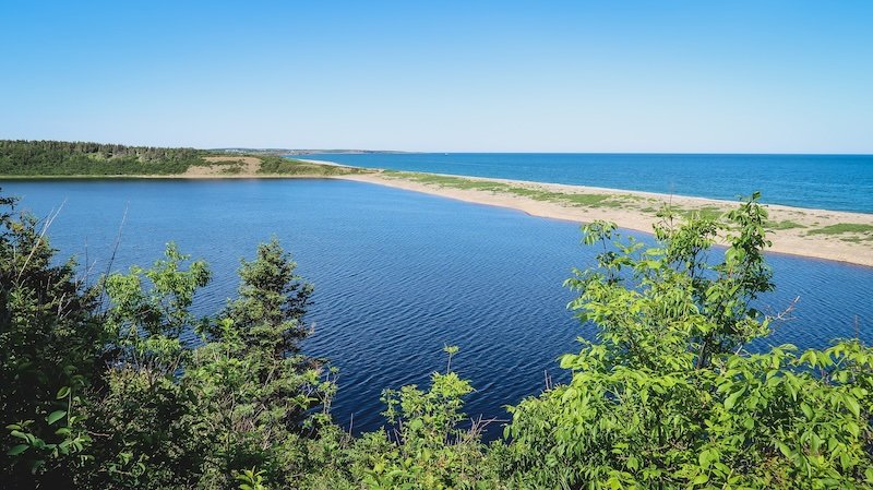 A sweeping view from a high vantage point overlooking vivid blue waters, a narrow sandspit, and lush green forest meeting the Atlantic Ocean along the scenic Cabot Trail in Cape Breton Highlands National Park, Nova Scotia