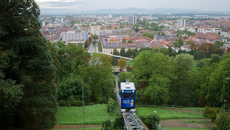 View over Freiburg im Breisgau, Germany, from the Schlossberg funicular railway, showing the car ascending through lush green trees toward the hilltop lookout with the cityscape stretching out below