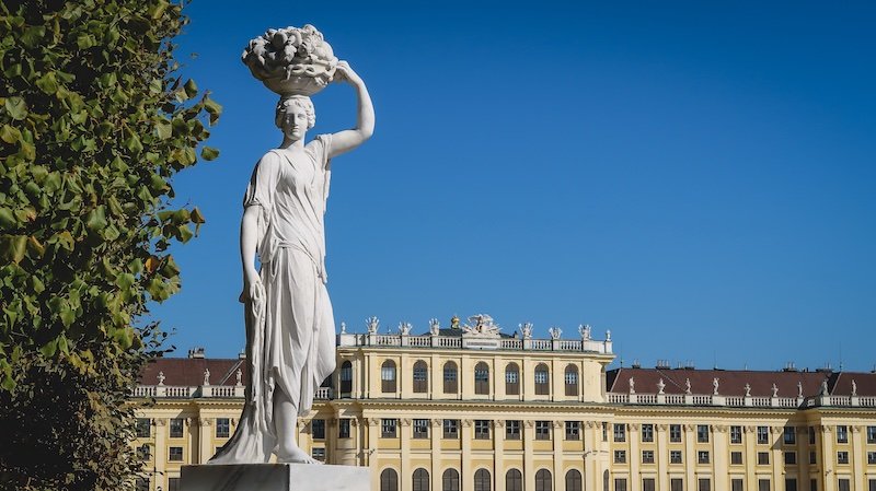 Elegant marble statue of a woman holding a fruit basket in the gardens of Schönbrunn Palace, with the grand yellow baroque façade of the palace in the background under a clear blue sky in Vienna, Austria Elegant marble statue of a woman holding a fruit basket in the gardens of Schönbrunn Palace, with the grand yellow baroque façade of the palace in the background under a clear blue sky in Vienna, Austria