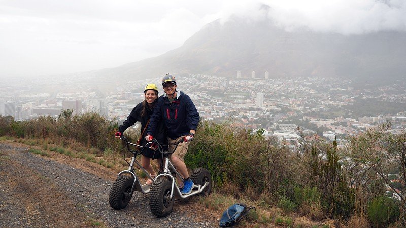 Nomadic Samuel and Audrey Bergner from That Backpacker riding rugged off-road scooters with Scootours on a misty downhill trail overlooking Cape Town, with fog rolling over Table Mountain in the background