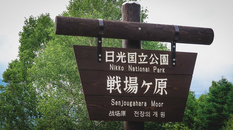 Wooden entrance sign for Senjogahara Moor in Nikko National Park, Nikko, Japan, with bilingual text in Japanese, English, Chinese, and Korean set against a backdrop of lush green trees
