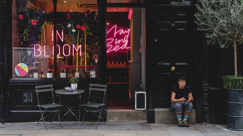 Street view of a trendy boutique in London’s Seven Dials, featuring neon signs reading “In Bloom” and “Stay in Bed,” with a small café table outside and a passerby seated nearby, showcasing the neighborhood’s vibrant and creative shopping scene
