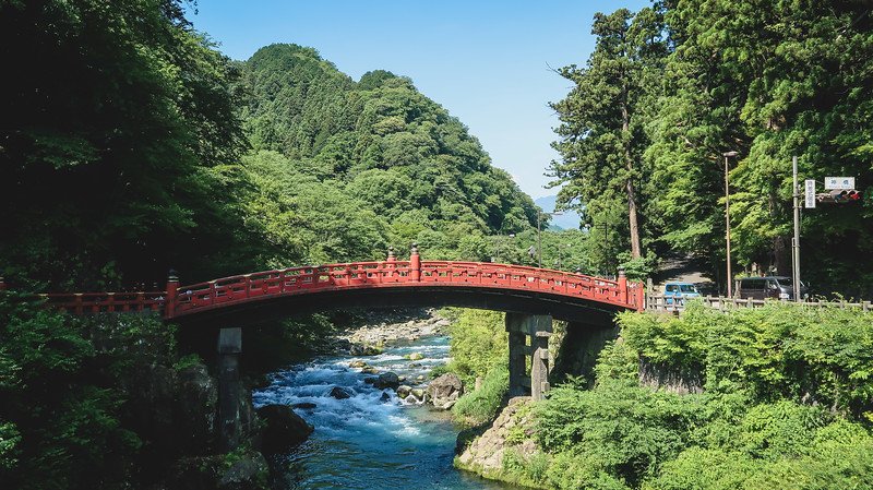 The iconic vermilion-lacquered Shinkyo Bridge spanning the crystal-clear Daiya River in Nikko, Japan, surrounded by lush green forested mountains under a bright blue summer sky