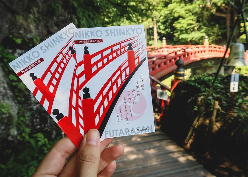 Close-up of two colorful entry tickets for Shinkyo Bridge in Nikko, Japan, held up in front of the iconic vermilion-lacquered sacred bridge surrounded by lush green forest on a sunny day