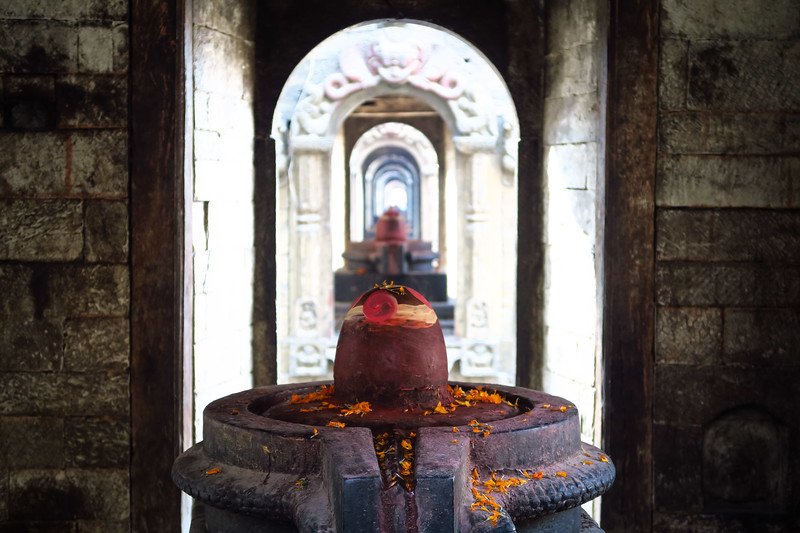 Hindu Shiva lingam shrine inside Pashupatinath Temple in Kathmandu, Nepal, framed by a sacred corridor of stone doorways adorned with marigold offerings and ritual flowers
