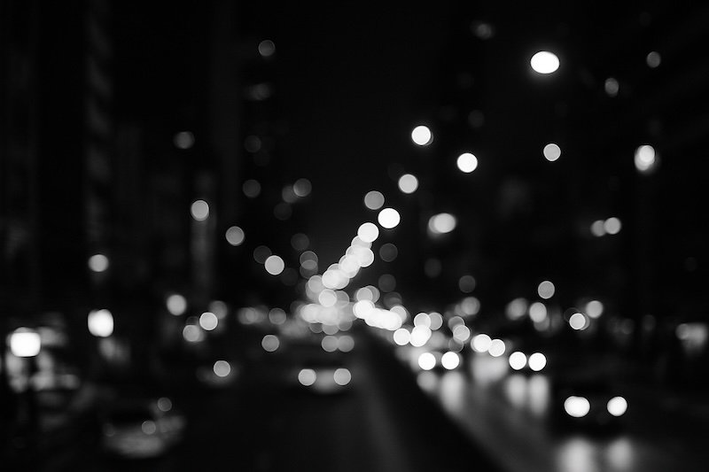 A dreamy black-and-white nighttime photograph of Silom Road in Bangkok, Thailand, captured with a shallow depth of field that turns city lights and traffic into soft circular bokeh, evoking the vibrant yet atmospheric energy of the urban night