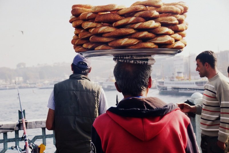 Street vendor carrying a towering tray of golden sesame-covered simit balanced on his head while walking along the waterfront near the Galata Bridge in Istanbul, Turkey, with fishermen and ferries in the background