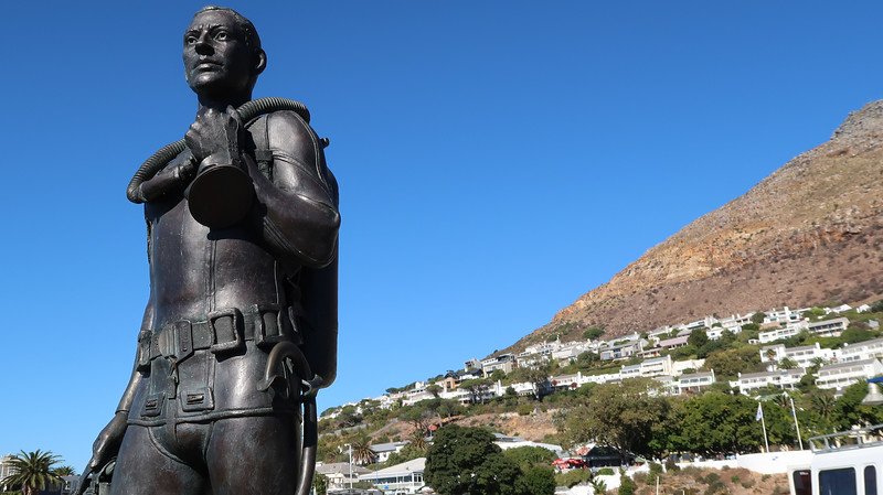 Close-up view of the bronze statue of a diver in Simon’s Town, South Africa, standing proudly against a vivid blue sky with the mountainside and white houses in the background, photographed during a scenic Cape Peninsula sidecar tour from Cape Town