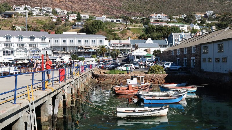 Scenic view of Simon’s Town harbour in South Africa, with small fishing boats bobbing in the water, a lively pier with visitors, and seafood restaurants lining the waterfront, captured during a Cape Peninsula sidecar tour from Cape Town