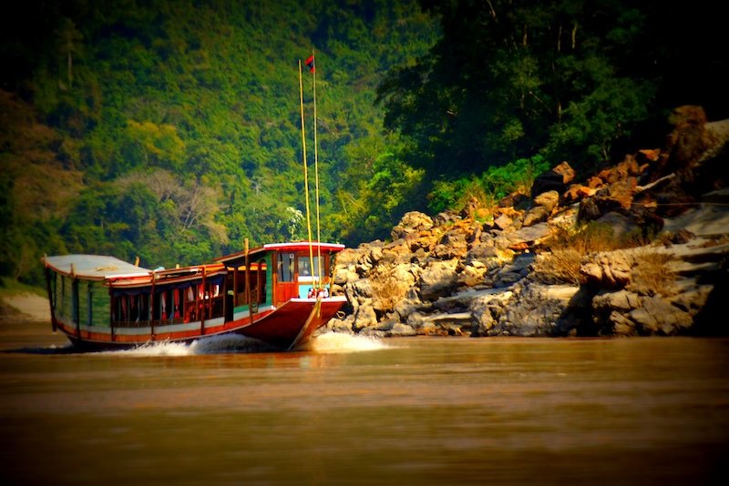A traditional slow boat glides along the muddy waters of the Mekong River in Laos, passing rocky riverbanks and lush green jungle. This iconic vessel is a popular way to travel between Thailand and Luang Prabang, offering scenic views and cultural immersion