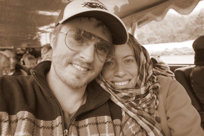 Nomadic Samuel and Audrey Bergner of That Backpacker smiling and bundled up during their journey on the slow boat to Laos. This sepia-toned travel portrait captures the cozy, adventurous atmosphere aboard the Mekong River vessel en route to Luang Prabang