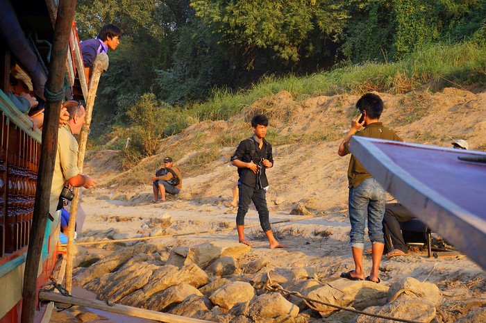 Passengers on the slow boat to Laos watch as young locals gather on the rocky riverbank at sunset. One man chats on his phone while another stands barefoot, capturing an authentic moment during the Mekong River journey toward Luang Prabang