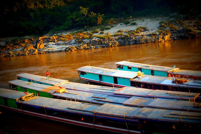 A row of traditional slow boats moored along the muddy banks of the Mekong River in Laos, bathed in warm golden afternoon light. The vibrant turquoise and red wooden boats contrast beautifully with the earthy river tones and rocky shoreline