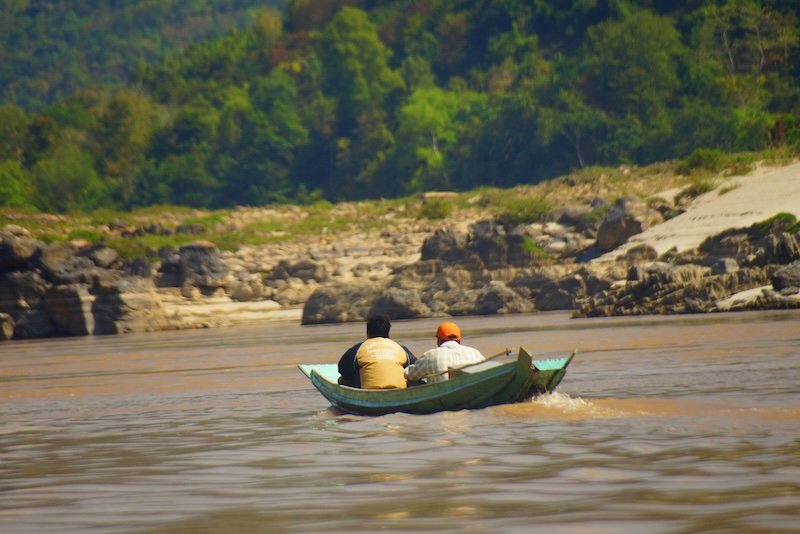 Two men travel upstream in a small wooden motorboat on the muddy waters of the Mekong River in Laos, surrounded by rocky riverbanks and dense jungle. This scene captures the daily river life and transportation used along this iconic Southeast Asian waterway