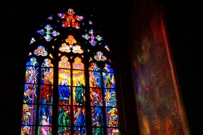 Vibrant Art Nouveau stained glass window inside St. Vitus Cathedral at Prague Castle in Prague, Czech Republic, depicting biblical figures with colorful light streaming onto the cathedral wall.
